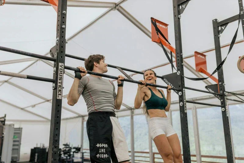 Man and woman performing pull-ups on a black metal rig, smiling during their workout.