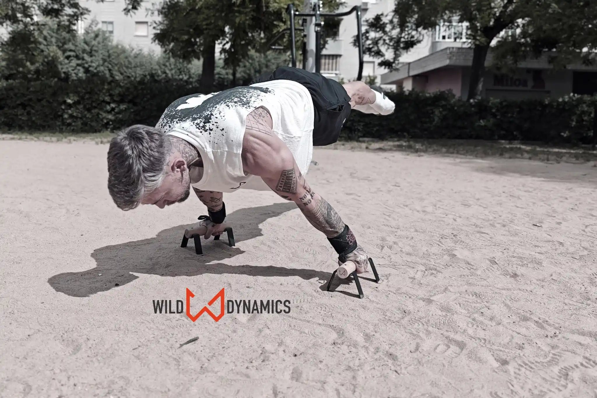 Man performing a calisthenics push-up on black and wood parallettes in a sandy park.