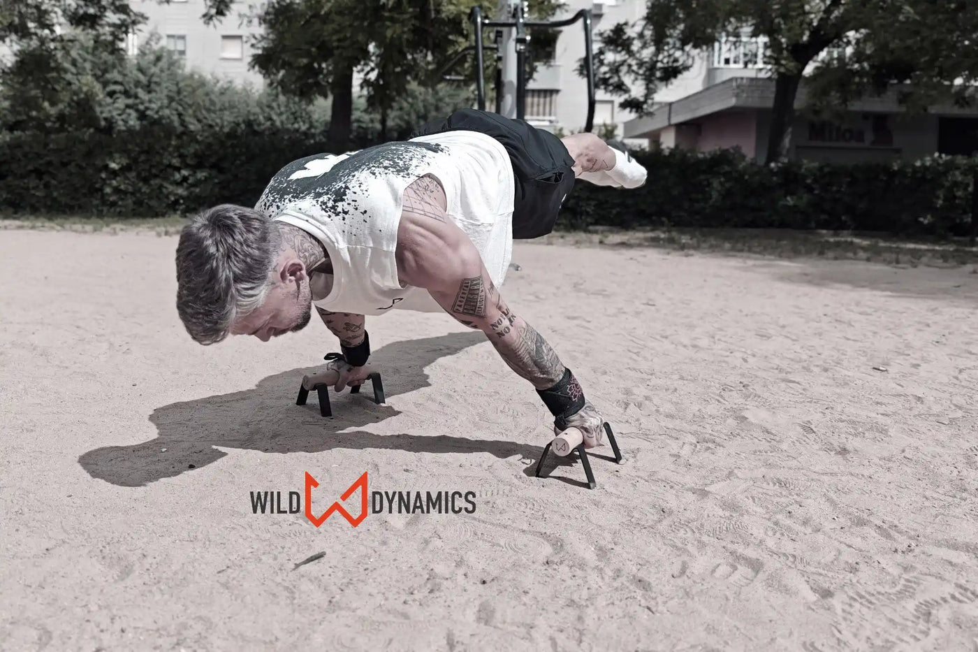 Man performing a calisthenics push-up on black and wood parallettes in a sandy park.