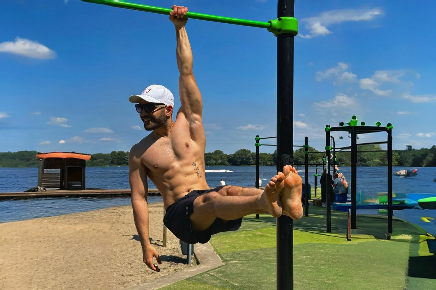 Man in sunglasses and hat performing a one-arm hang on a green bar at an outdoor calisthenics park.