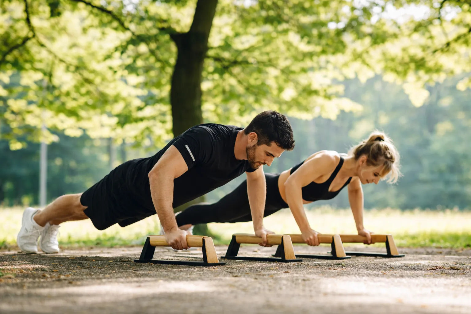 Man and woman doing push-ups on wooden parallettes outdoors, demonstrating beginner skills.