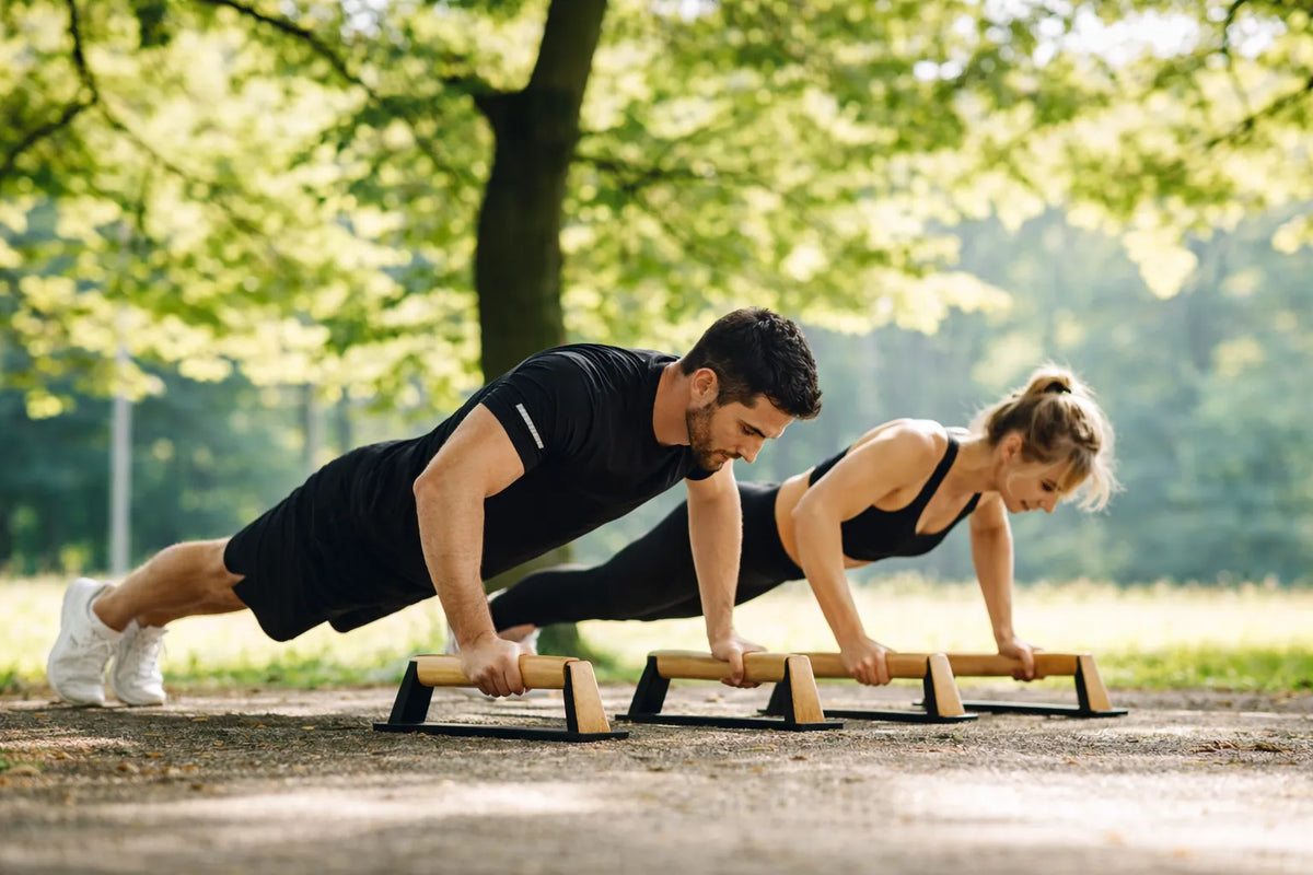 Man and woman doing push-ups on wooden parallettes outdoors, demonstrating beginner skills.