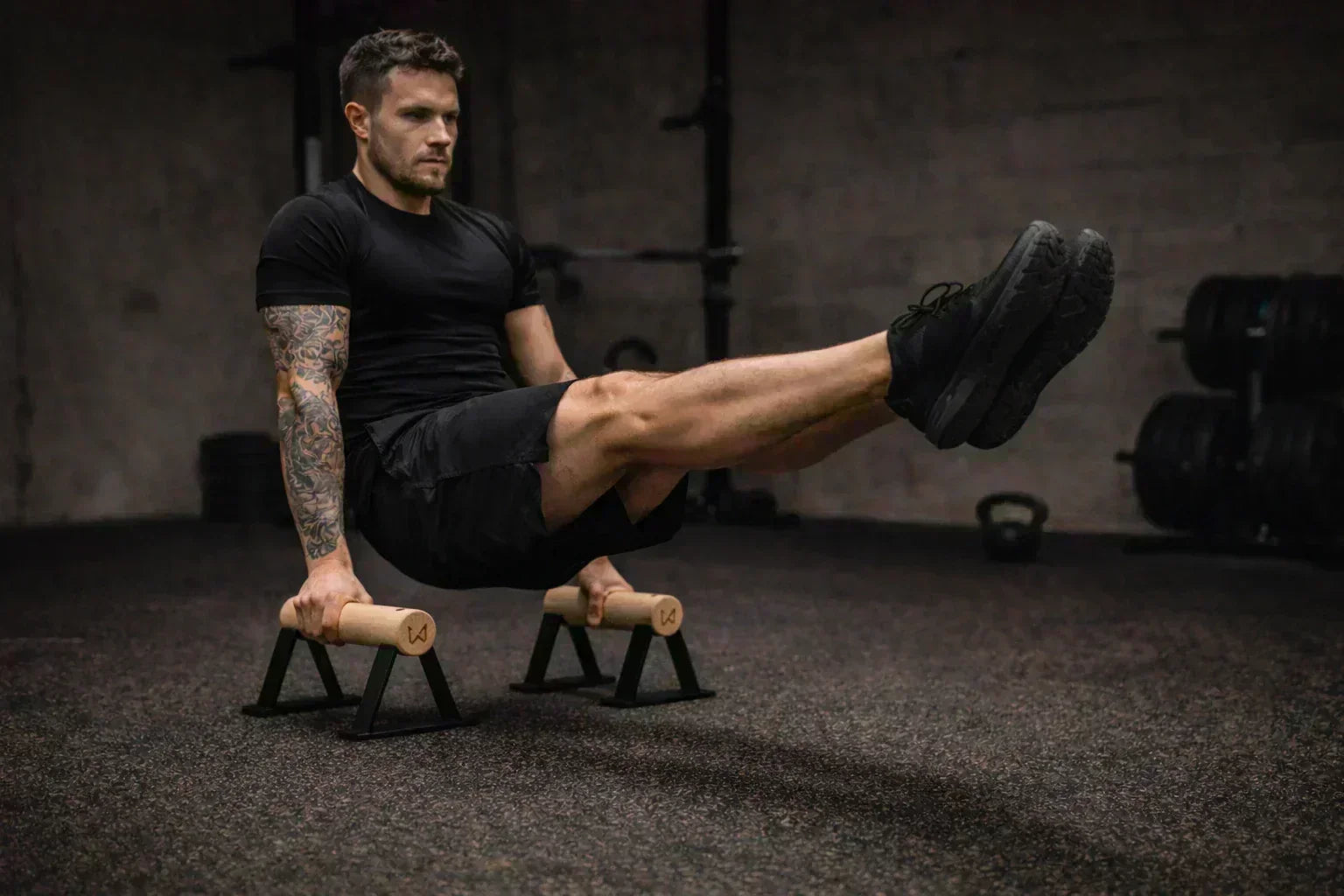 Man in black activewear performing an L-sit on wooden parallettes in a gym.