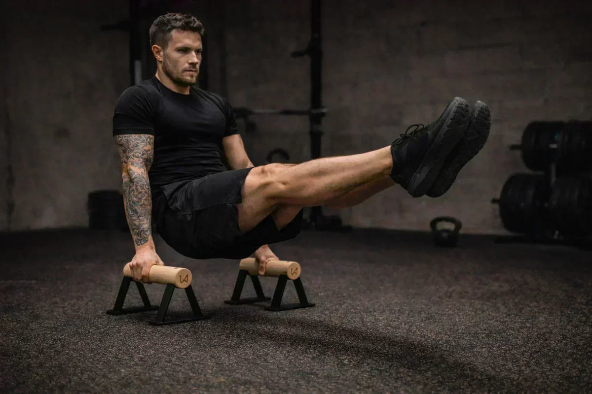 Man in black activewear performing an L-sit on wooden parallettes in a gym.