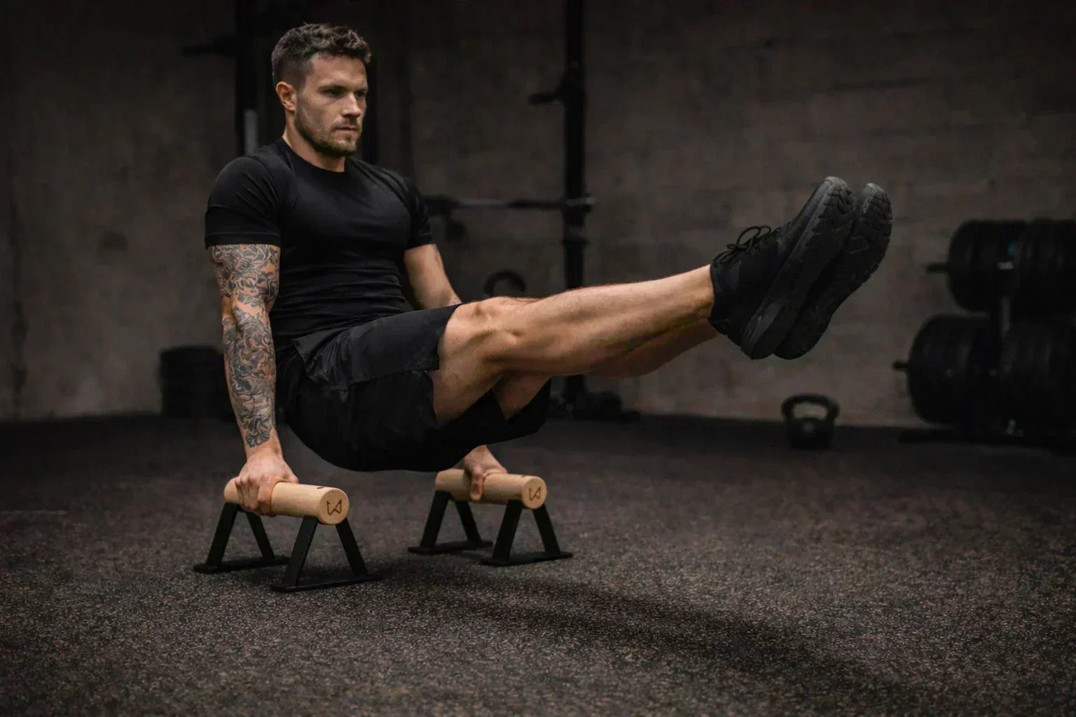 Man in black activewear performing an L-sit on wooden parallettes in a gym.
