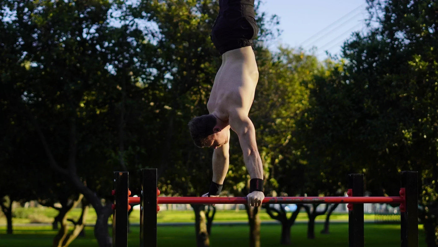 Muscular man performing a handstand on red parallettes outdoors, demonstrating advanced strength.