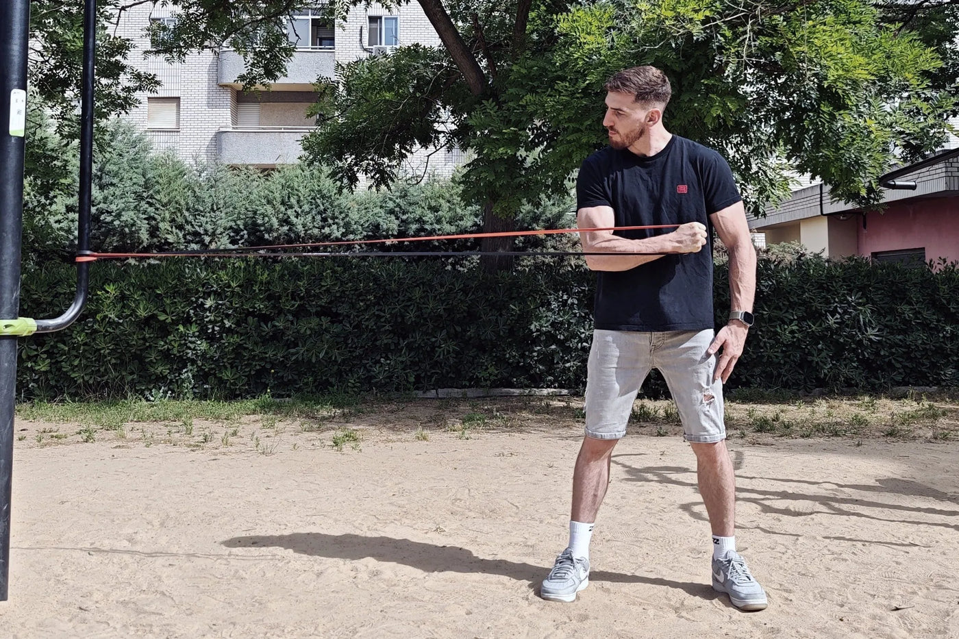 Man in black t-shirt and grey shorts doing a calisthenics warm-up with a resistance band outdoors.
