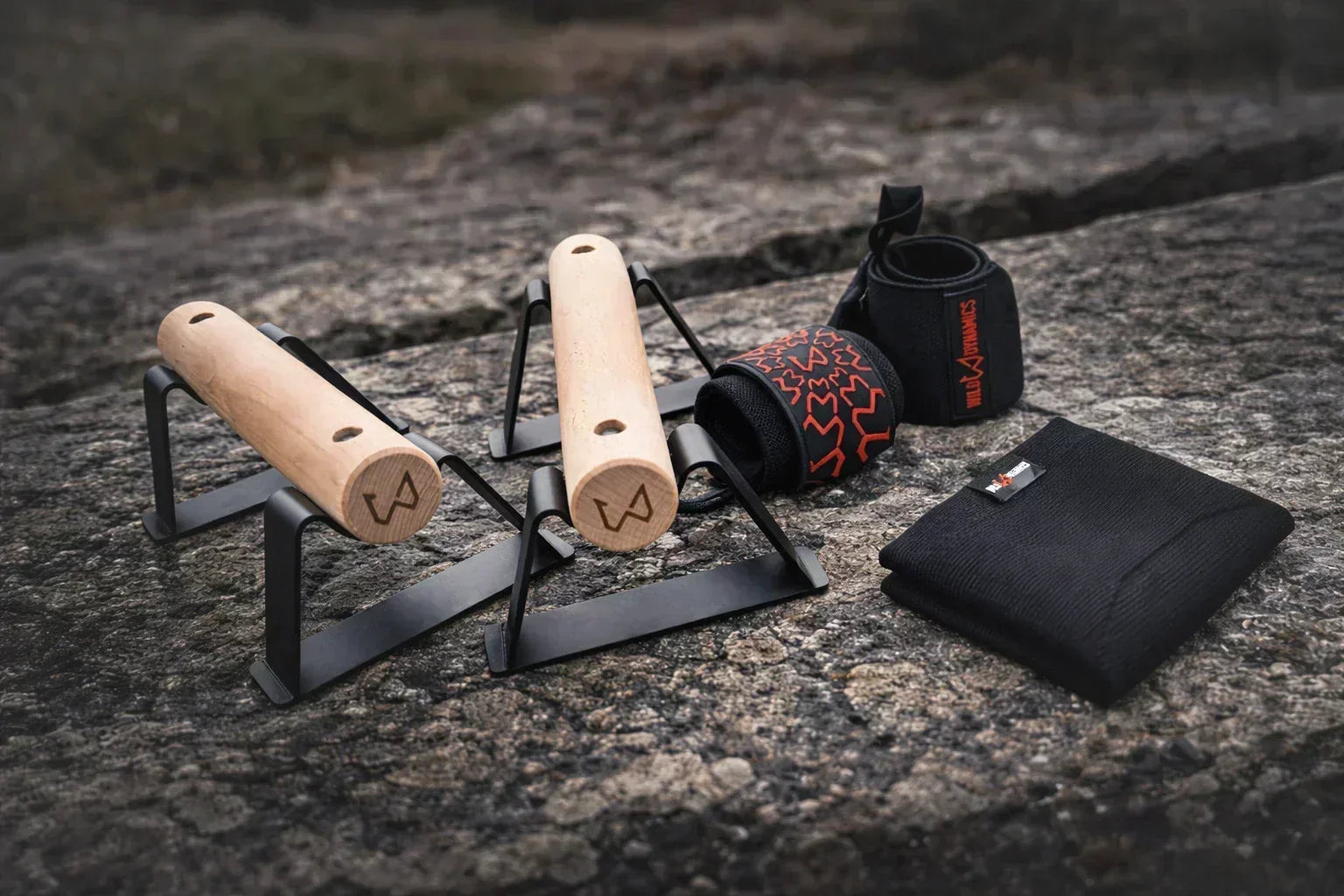 Portable calisthenics equipment: wooden push-up bars, black wrist wraps, and a folded mat on a rocky surface.