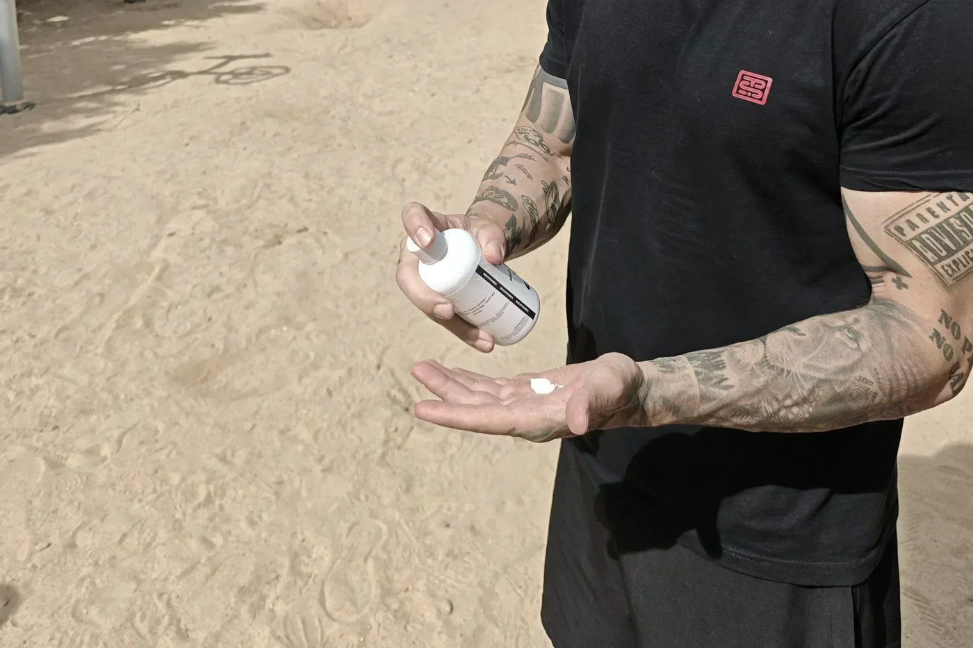 Man with tattooed arms applying white lotion from a bottle to his hand outdoors.