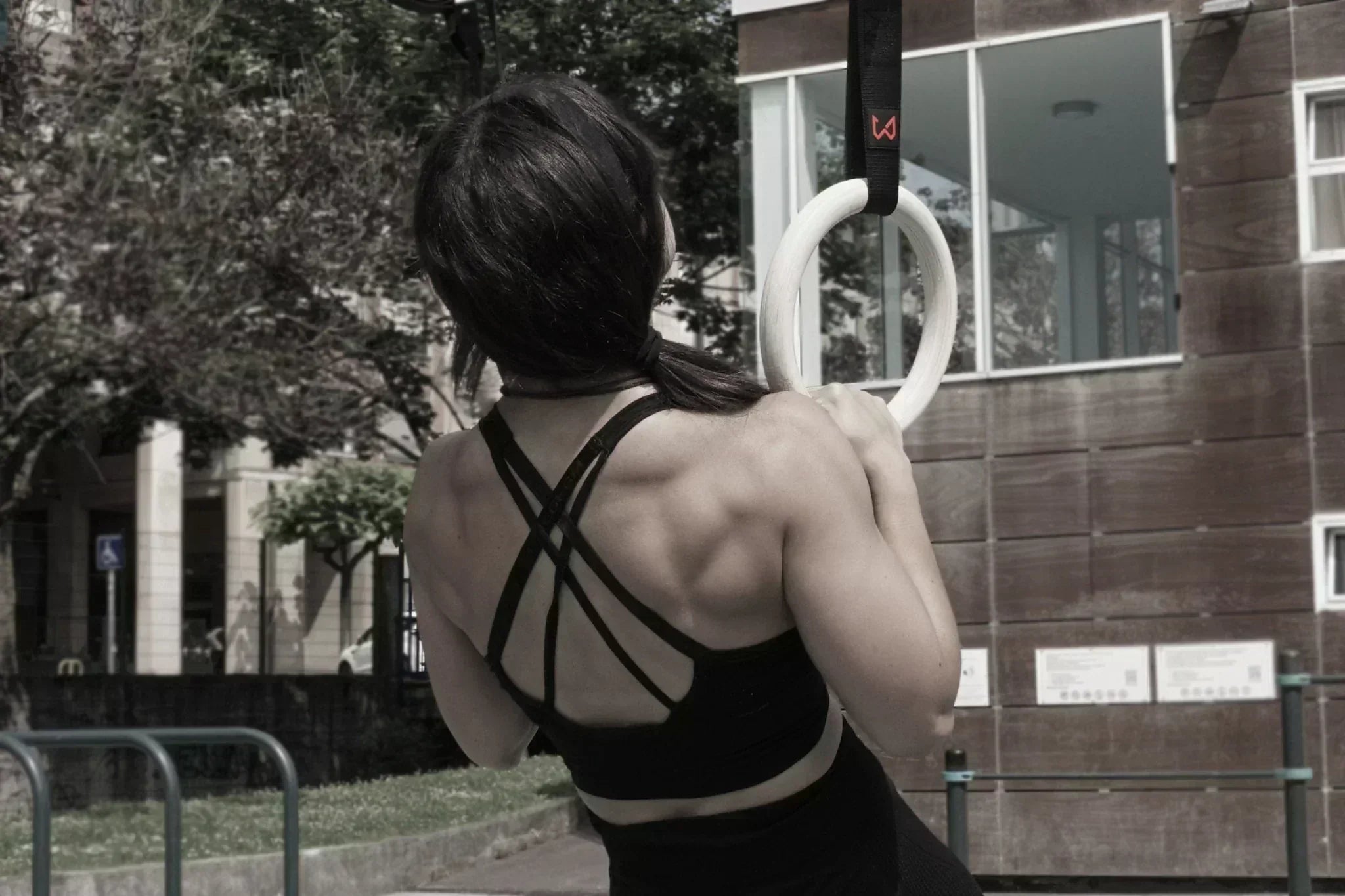 Woman in a black sports bra performing a calisthenics exercise on white wooden gymnastic rings outdoors.