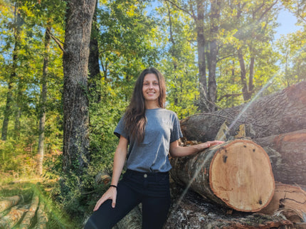 Woman standing next to a large log in a forest wearing gray organic cotton tshirt of wild dynamics