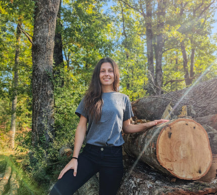 Woman standing next to a large log in a forest wearing gray organic cotton tshirt of wild dynamics