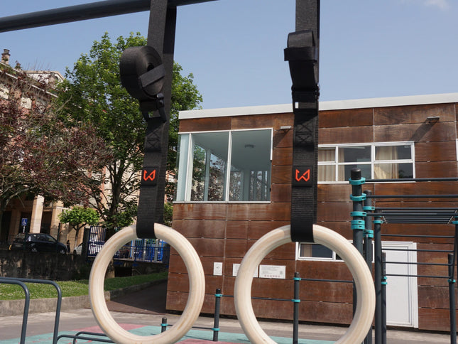 Pair of light birch wood gymnastics rings with black straps hanging from a pull-up bar outdoors.