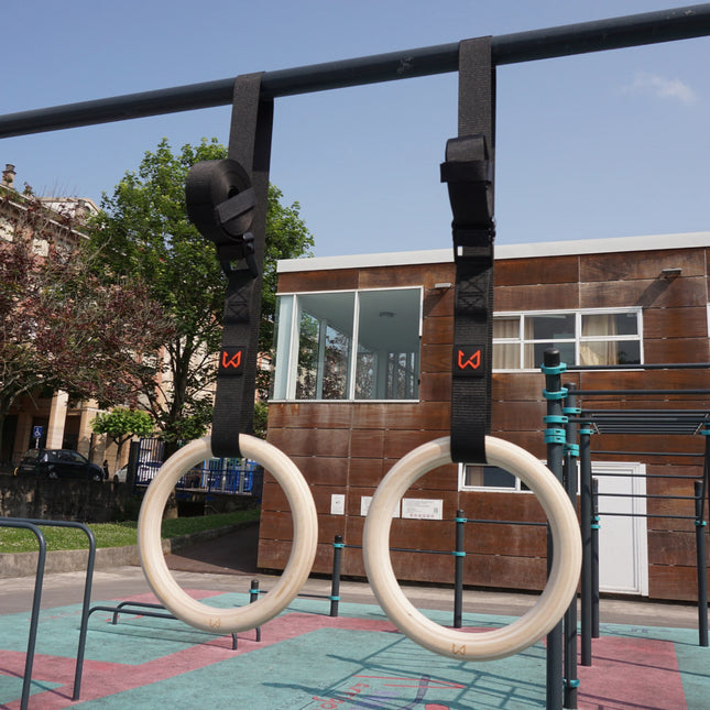 Pair of light birch wood gymnastics rings with black straps hanging from a pull-up bar outdoors.