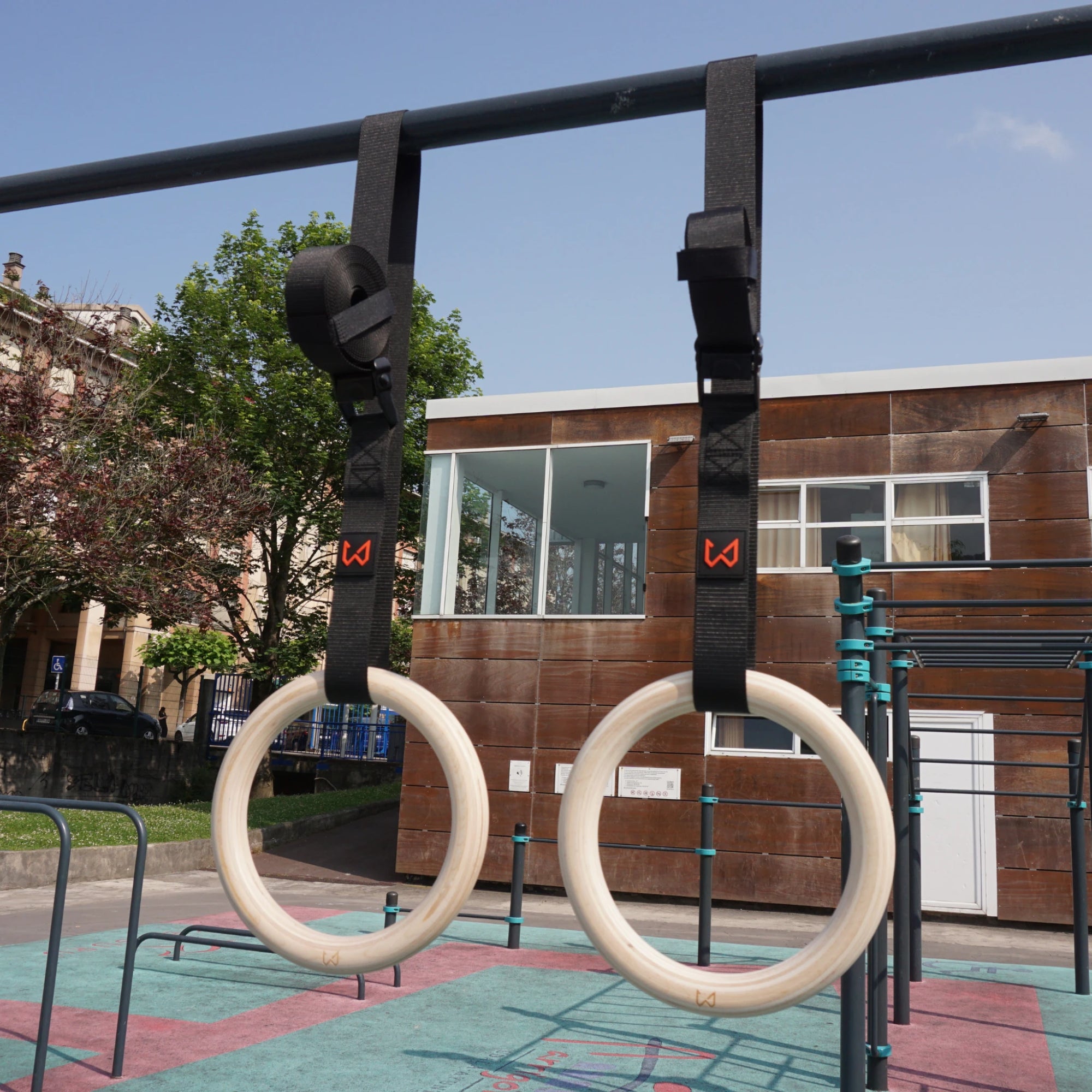 Pair of light birch wood gymnastics rings with black straps hanging from a pull-up bar outdoors.