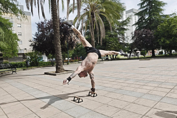Person performing a one arm handstand on parallettes in a park.